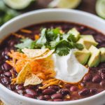 A bowl of black bean soup served with sour cream, shredded cheese, diced avocado