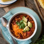 A bowl of chili garnished with shredded cheese, diced avocado, and fresh cilantro