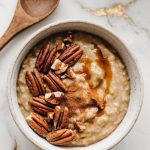 A bowl of freshly served pumpkin spice oatmeal, topped with chopped pecans, maple syrup