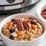 A bowl of steaming carrot cake oatmeal served with toppings like pecans, raisins