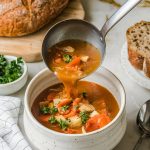 A ladle pouring soup into a white bowl, surrounded by a rustic loaf of bread, a small bowl of parsley, and a spoon