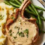 tender pork chops plated with creamy gravy, sprinkled with parsley, served alongside mashed potatoes and green beans