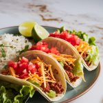 A plate with freshly assembled tacos in soft tortillas, topped with lettuce, diced tomatoes