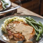 A plate of tender pork chops drizzled with creamy dairy-free gravy, served with mashed potatoes and green beans