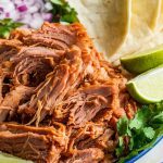 Plate of shredded, crispy carnitas served with tortillas, lime wedges, diced onions, fresh cilantro