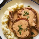 Juicy pork chops plated with creamy sauce poured over, garnished with parsley, and served with cauliflower mash on the side