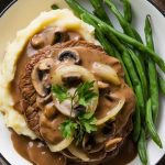 A serving of Salisbury steak plated with creamy mashed potatoes and buttered green beans