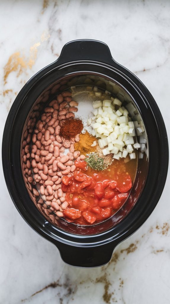 A crockpot filled with pinto beans, water, diced onions, garlic, spices, and canned tomatoes