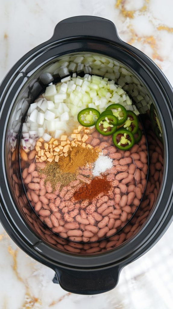 A crockpot filled with rinsed pinto beans, water, diced onion, minced garlic, sliced jalapeño, and spices like cumin and paprika sprinkled on top