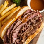 a French dip sandwich served on a wooden board, with a small bowl of au jus on the side