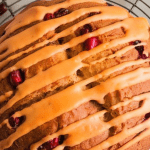 A freshly baked loaf of cranberry orange bread being drizzled with orange glaze