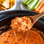 A wooden spoon lifting a scoop of creamy buffalo chicken dip from the crockpot, cheese stretching slightly