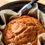 Freshly cooked banana bread being lifted from the crockpot using parchment paper