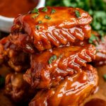 A rustic wooden plate piled high with honey BBQ chicken wings, glistening with a thick, caramelized glaze