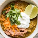 a hearty bowl of slow cooker pork chili verde being finished with a sprinkle of chopped fresh cilantro, a dollop of sour cream, shredded cheese, and a lime wedge on the side