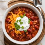 a steaming bowl of finished Crockpot Beer Chili, thick and chunky with beef, beans, and tomatoes, topped with melted shredded cheese, a swirl of sour cream, and fresh chopped green onions
