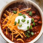 a finished bowl of chili con carne, with melty shredded cheddar, a dollop of sour cream, and chopped green onions on top