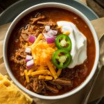 a steaming bowl of shredded beef Texas chili topped with shredded cheddar cheese, diced red onions, jalapeño slices, and a dollop of sour cream