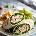 two sliced chicken Caesar wraps on a white plate, showing creamy filling and fresh lettuce, served with chips and grapes