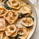 a bowl of creamy tortellini Alfredo, tortellini coated in thick sauce, bits of spinach throughout, sprinkled with extra parmesan and red pepper flakes