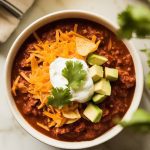 a bowl of thick chili topped with melty cheddar cheese, a dollop of sour cream, diced avocado, and crushed tortilla chips