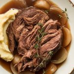 the shredded pot roast surrounded by caramelized onions and broth, steam rising, served on a plate with a side of mashed potatoes
