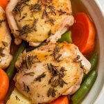 a white bowl filled with steaming herb-roasted chicken thigh resting on tender carrots, potatoes, green beans