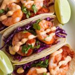 a taco being assembled: soft corn tortilla filled with saucy shrimp, crunchy purple slaw, creamy chipotle drizzle, and bright green cilantro