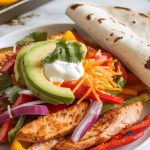 an open flour tortilla being filled with roasted chicken fajitas, topped with avocado slices, sour cream, and chopped cilantro