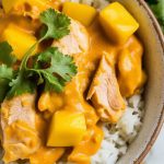 a serving bowl filled with fluffy white rice topped with creamy mango chicken curry, juicy chunks visible
