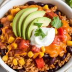 a burrito bowl being finished with creamy sour cream, shredded cheddar cheese, diced avocado, and chopped fresh cilantro on top of the hot chicken and rice mix