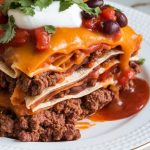 sliced crockpot Mexican lasagna, melty cheese on top, visible stacked layers of tortilla, beef, beans, and salsa, topped with fresh chopped cilantro and sour cream