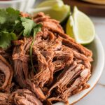 crispy shredded pork carnitas piled onto a white ceramic plate, garnished with fresh cilantro and lime wedges on the side, golden crispy edges visible on the pork