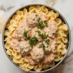 a scoop of creamy meatball stroganoff being served over a pile of cooked egg noodles, with fresh parsley sprinkled on top
