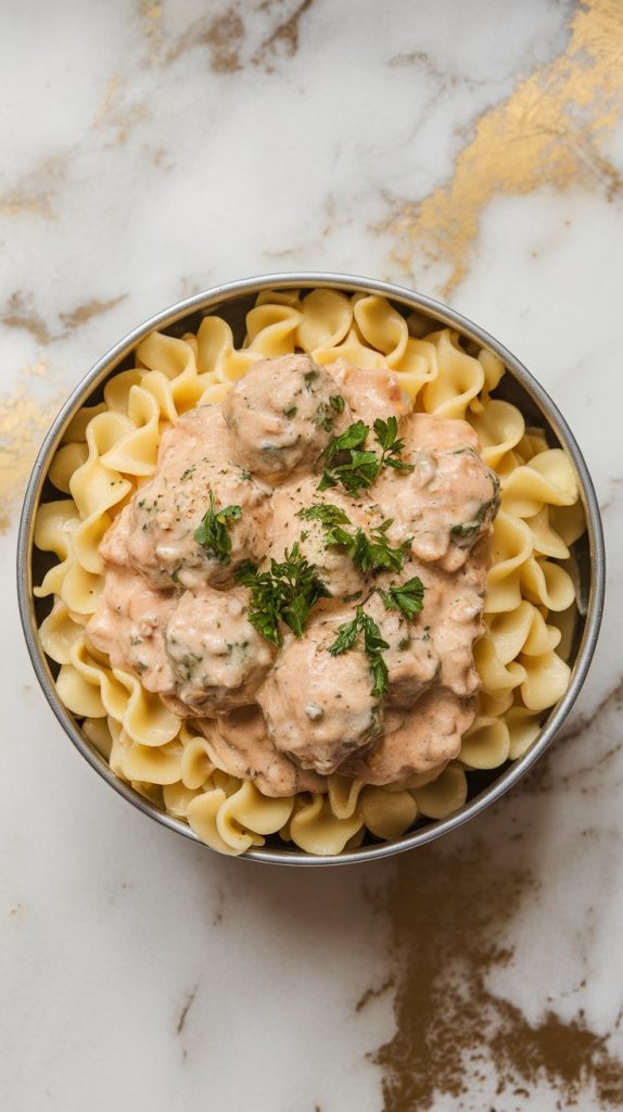 a scoop of creamy meatball stroganoff being served over a pile of cooked egg noodles, with fresh parsley sprinkled on top