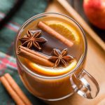 a steaming mug of crockpot apple cider with cinnamon sticks, floating orange slices, and star anise in rich golden brown liquid, served in a clear glass mug on a wood tray