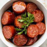 finished kielbasa bites being spooned into a white bowl, garnished with chopped parsley