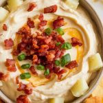 a ceramic bowl filled with warm loaded baked potato dip, showing a smooth, slightly runny off-white base with a faint yellow tint