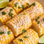 creamy corn on the cob in a white bowl, topped with chopped parsley and drizzled with coconut sauce, lime wedges on the side