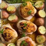 fully cooked lemon garlic chicken sheet pan fresh out of the oven, featuring exactly 4 golden, crispy-skinned bone-in chicken thighs nestled among roasted halved baby potatoes arranged cut-side up and tightly packed