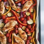 a parchment-lined sheet pan just pulled from the oven, showing a slightly crowded, naturally scattered mixture of roasted chicken breast strips with golden edges, roasted red and yellow bell peppers, zucchini half-moons, red onion slices