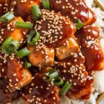finished General Tso’s Chicken being garnished with sesame seeds and chopped green onions, glossy sauce shining, fluffy white rice on the side